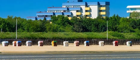 Un edificio costero con varios balcones, una playa arenosa al frente y un cielo azul despejado.