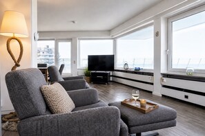 A modern living room with grey sofas, a flat-screen TV, and a large window with a view of the city. - Luxurious penthouse Duhner Strand with sea view | Haus Hanseatic (Cuxhaven)