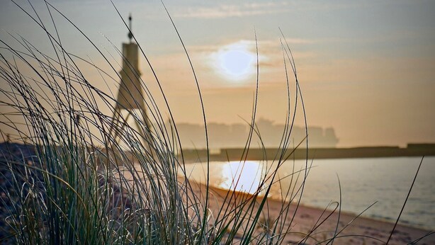 En strand med højt græs i forgrunden og et fjerntliggende bybillede.