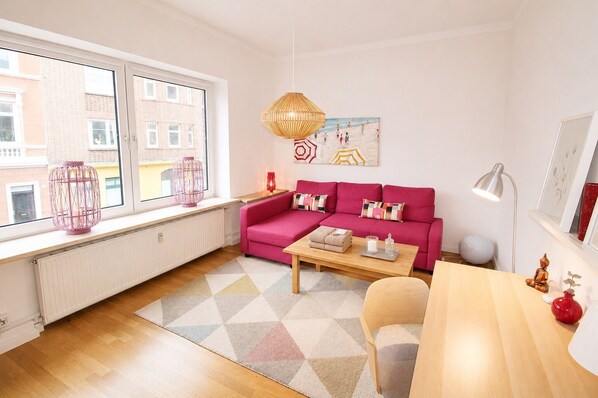 Een moderne woonkamer met een roze bank, een houten salontafel en een uitzicht op gebouwen door grote ramen.