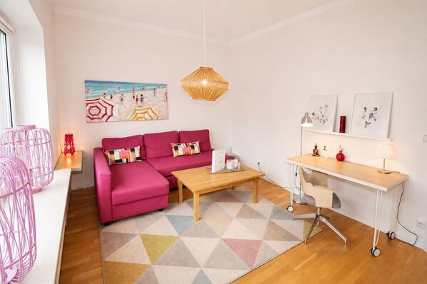Un salon moderne avec un canapé rose, une table basse en bois, un bureau avec une lampe et un tableau sur le mur représentant un paysage de plage.