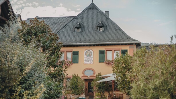 A building with a steeply pitched roof, a central entrance with a door, and a sign above the door. There are outdoor tables and chairs in front of the building. - KultUrlaub Mosel GbR (Kröv)