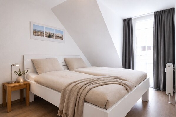 A modern bedroom with a white bed, a bedside table with a lamp, a picture on the wall, and a window with curtains.