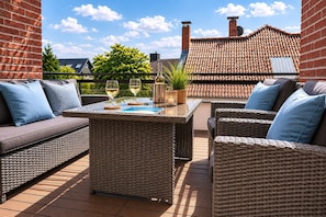 Um terraço no telhado com móveis de vime, uma mesa com copos e vista para edifícios e árvores.