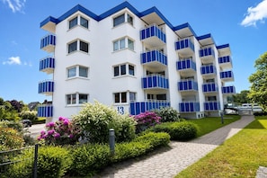 Un edificio residencial de varios pisos con balcones, un camino pavimentado y jardines bien cuidados.