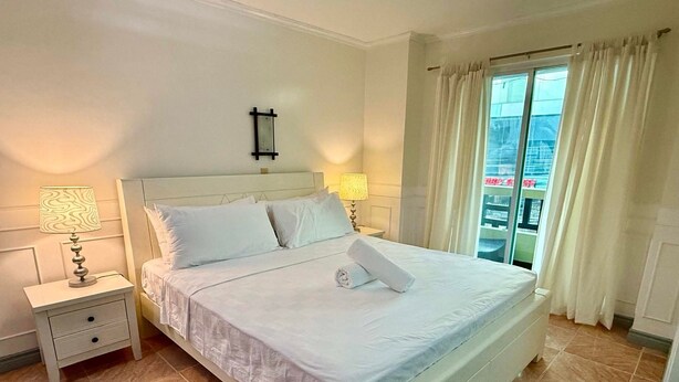 A neatly made bed with white linens, two bedside lamps, a small bedside table with a drawer, and a window with curtains. - Boracay Paradise Hotel (Boracay Island)