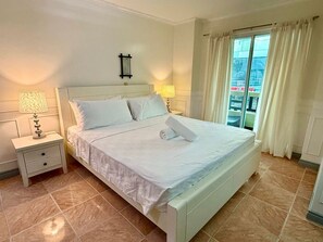 Een keurig opgemaakt bed met witte lakens, twee bedlampen, een klein nachtkastje met lade, en een raam met gordijnen.