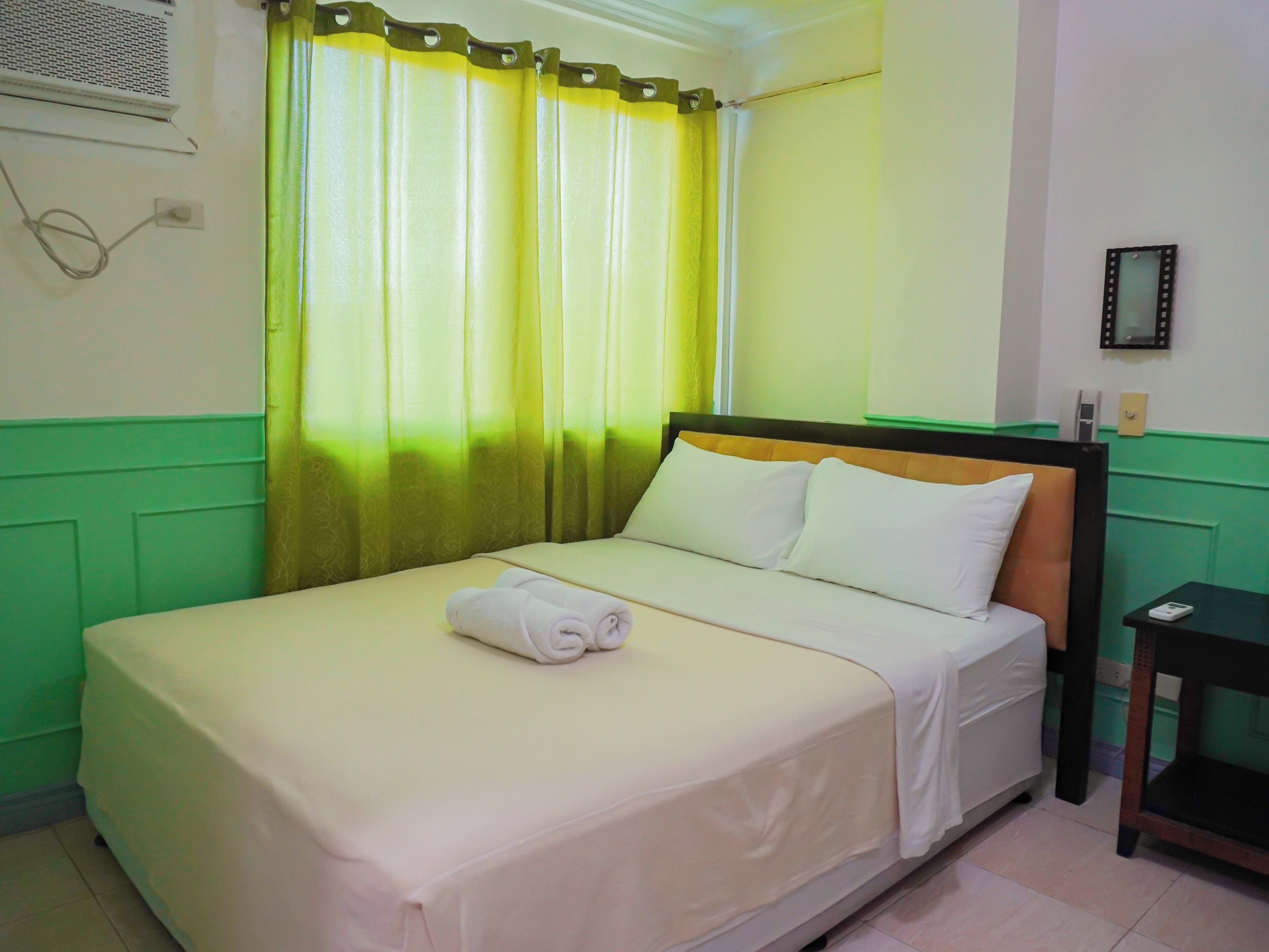 Habitación de hotel con cama, paredes verdes, ventana con cortinas amarillas y mesita de noche.