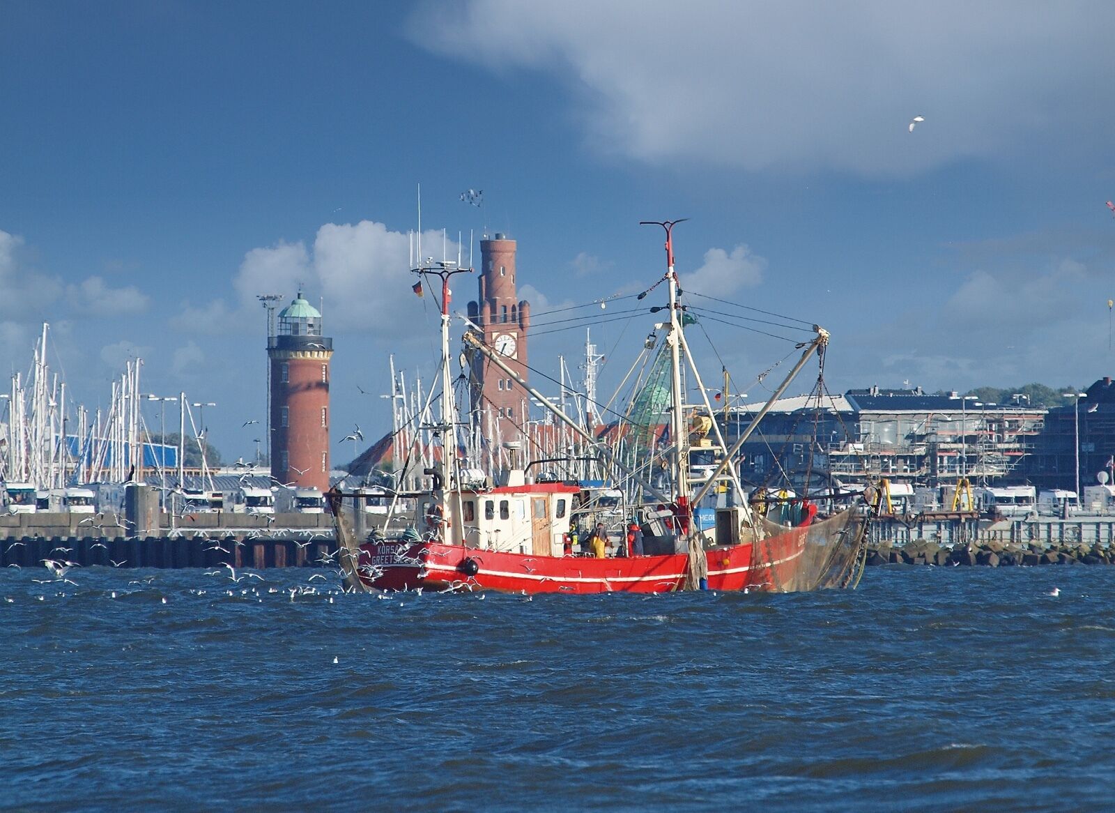 A harbor with a large fishing boat, a clock tower, and a lighthouse in the background.