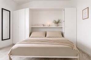 Un dormitorio con cama, armario, estante con libros y una vela, además de una cita enmarcada en la pared.