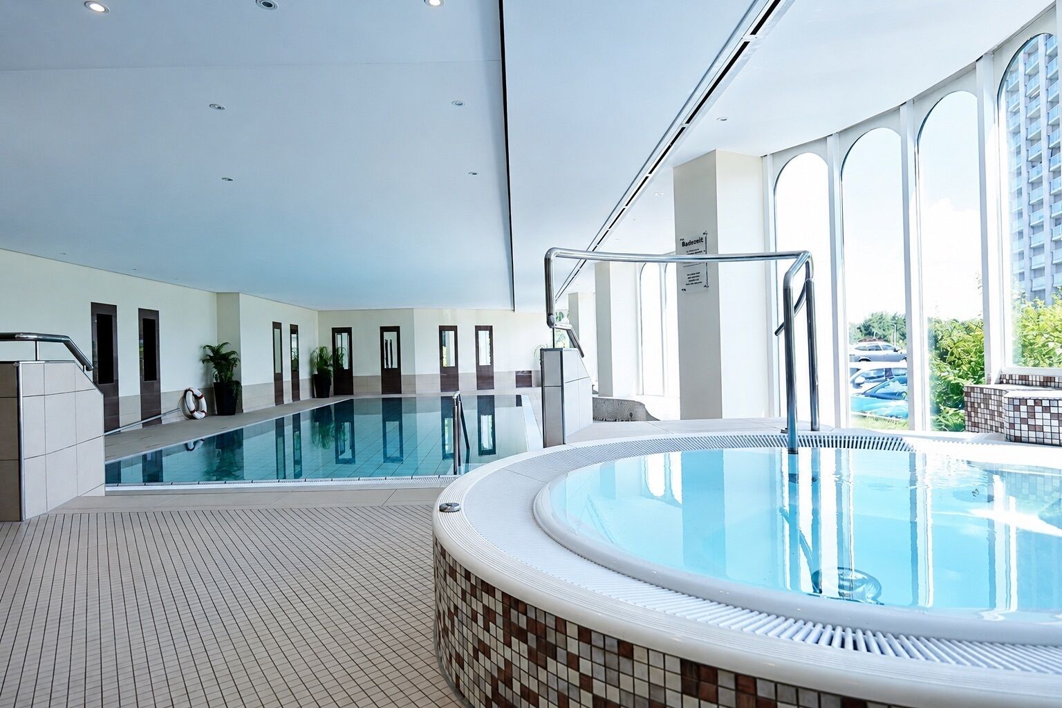 Une piscine intérieure moderne, dotée d’un espace séparé avec un jacuzzi, de grandes fenêtres et d’un sol carrelé.