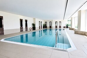 Una amplia piscina interior con paredes y suelo de baldosas, rodeada de ventanas y puertas.
