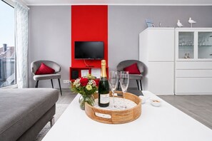 A modern living room with a red accent wall, a flat-screen TV, two grey armchairs, a white coffee table with a bottle of champagne and glasses, and a cabinet with glassware. - Vacation apartment near the beach in the heart of Duhnen with balcony & underground parking | Möwe (Cuxhaven)