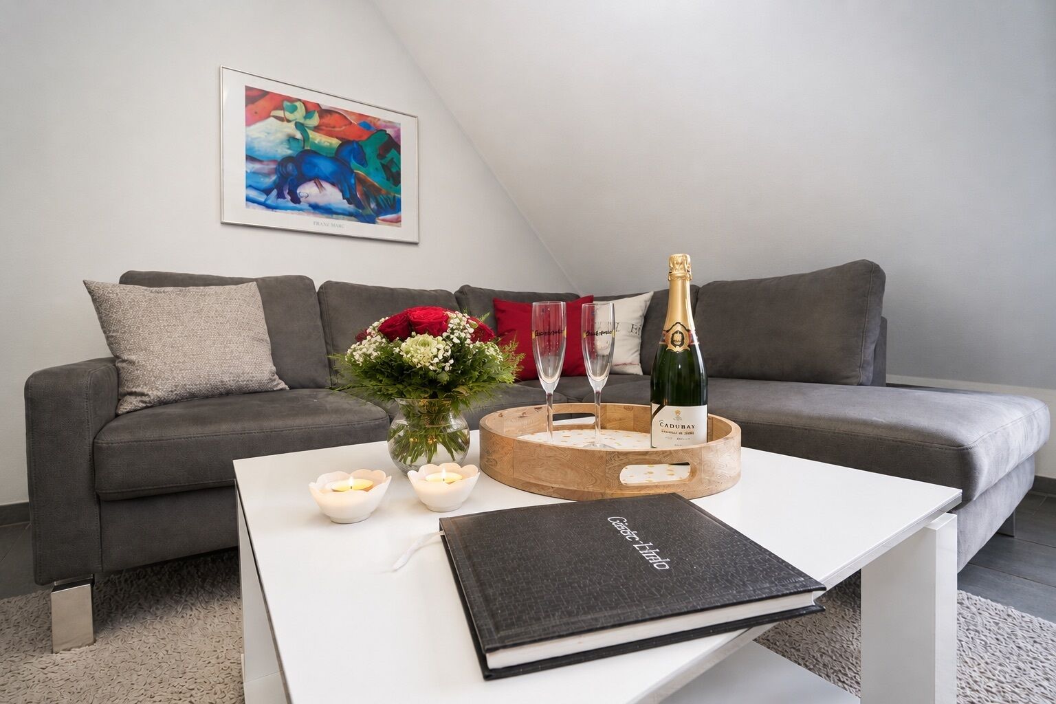En moderne stue med en grå sofa, et hvidt sofabord, et indrammet abstrakt maleri, en blomsterbuket, to vinglas, en champagneflaske og en bog.