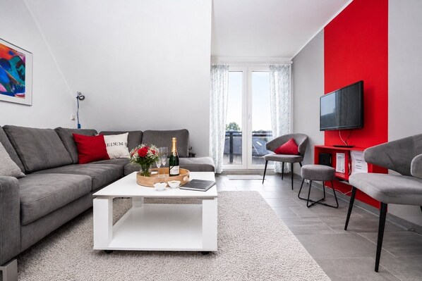 Ein modernes Wohnzimmer mit einem grauen Sofa, einem weißen Couchtisch, einer roten Akzentwand, einem Fernseher und Blick auf einen Balkon.
