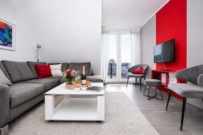 Ein modernes Wohnzimmer mit einem grauen Sofa, einem weiĂen Couchtisch, einer roten Akzentwand, einem Fernseher und Blick auf einen Balkon.