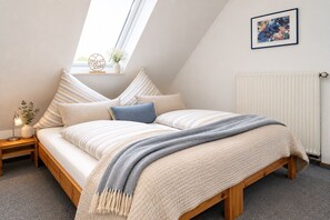 Ein Schlafzimmer mit schrÀgem Dach, einem Bett mit gestreifter BettwÀsche, einem Nachttisch mit Lampe, einem Fenster mit Ausblick und einer Heizung.