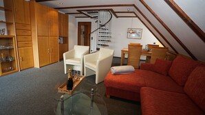 Un salon chaleureux de style loft, avec un canapé rouge, deux fauteuils blancs, une table basse en verre, des meubles en bois, un escalier en colimaçon et un coin repas avec des meubles en osier.