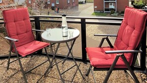 Un balcon avec deux chaises rouges et une petite table, donnant sur un quartier résidentiel avec des maisons et un ciel dégagé.