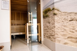 A sauna room with wooden benches and a glass door, a tiled floor, and a wall with a sand dune mural.