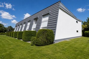 Un bâtiment moderne à l’extérieur blanc et au toit recouvert de tuiles grises, entouré de pelouses verdoyantes et de haies bien entretenues.