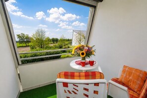 Varanda com vista para o verde e uma mesa com um vaso de flores e duas chávenas vermelhas.