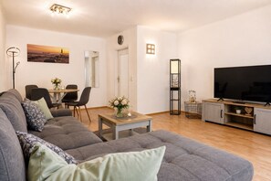 Ein Wohnzimmer mit grauem Sofa, einem Esstisch mit Stühlen, einem Fernseher und einem Gemälde an der Wand.
