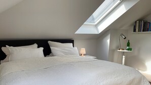 A neatly made bed with a white bedspread and pillows, a bedside lamp, and a small table with a bottle and glasses.