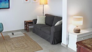 Ein Wohnzimmer mit einem Türkis-Armlehnstuhl, einem grauen Sofa, einem kleinen Holztisch, einer Lampe und einem Fernseher an der Wand.