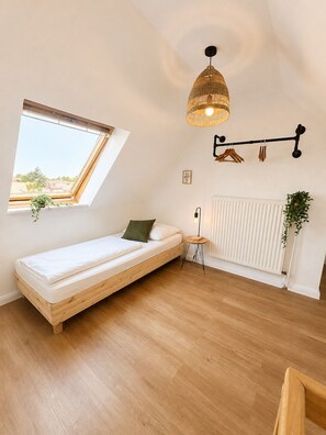 A small, single-room apartment with a slanted ceiling, a skylight, a bed, a radiator, a hanging light fixture, and a small table.