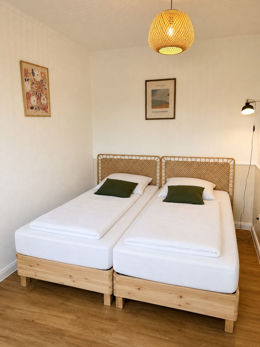 Um quarto com duas camas de madeira, roupa de cama branca, um candeeiro pendente e duas molduras com quadros na parede.