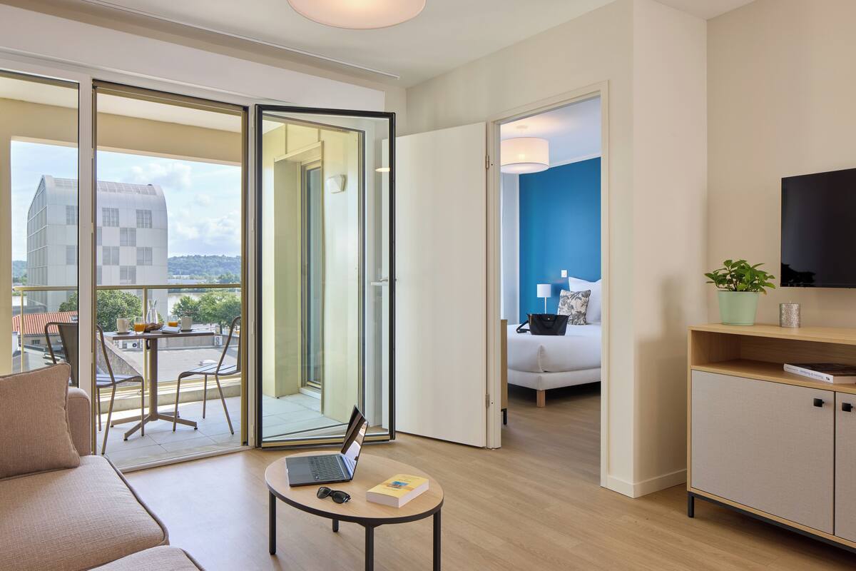 A modern hotel room with a balcony, a sofa, a small table with a laptop and book, and a TV on the wall.