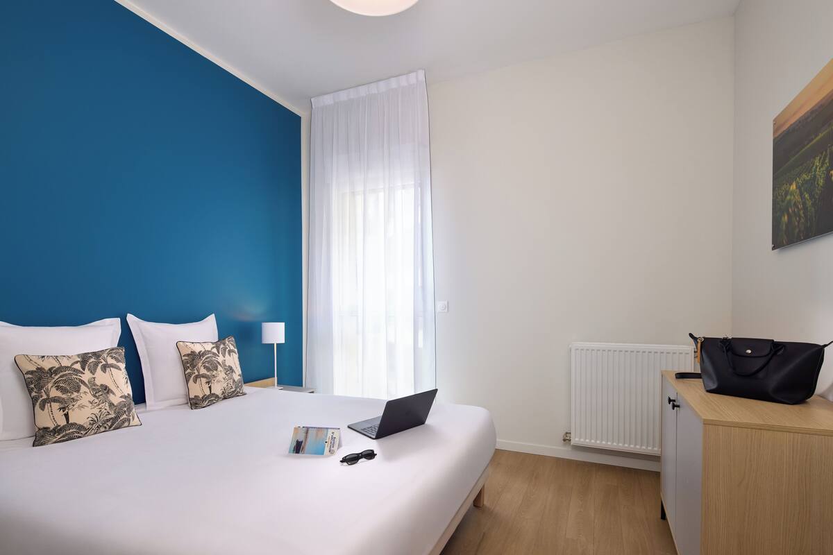 A hotel room with a bed, a laptop, a black handbag, and a white radiator.