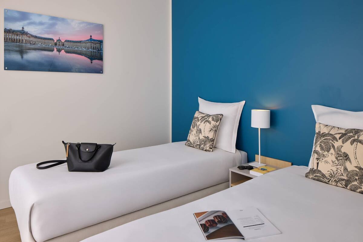 A hotel room with two beds, a nightstand, a lamp, a black handbag, and a framed picture on the wall.