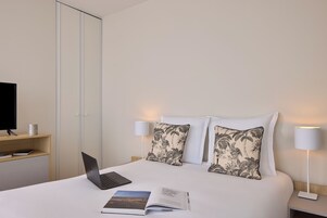 Une chambre d’hôtel avec un lit, un bureau avec un ordinateur portable, un livre, deux coussins décoratifs, une télévision et deux lampes de chevet.