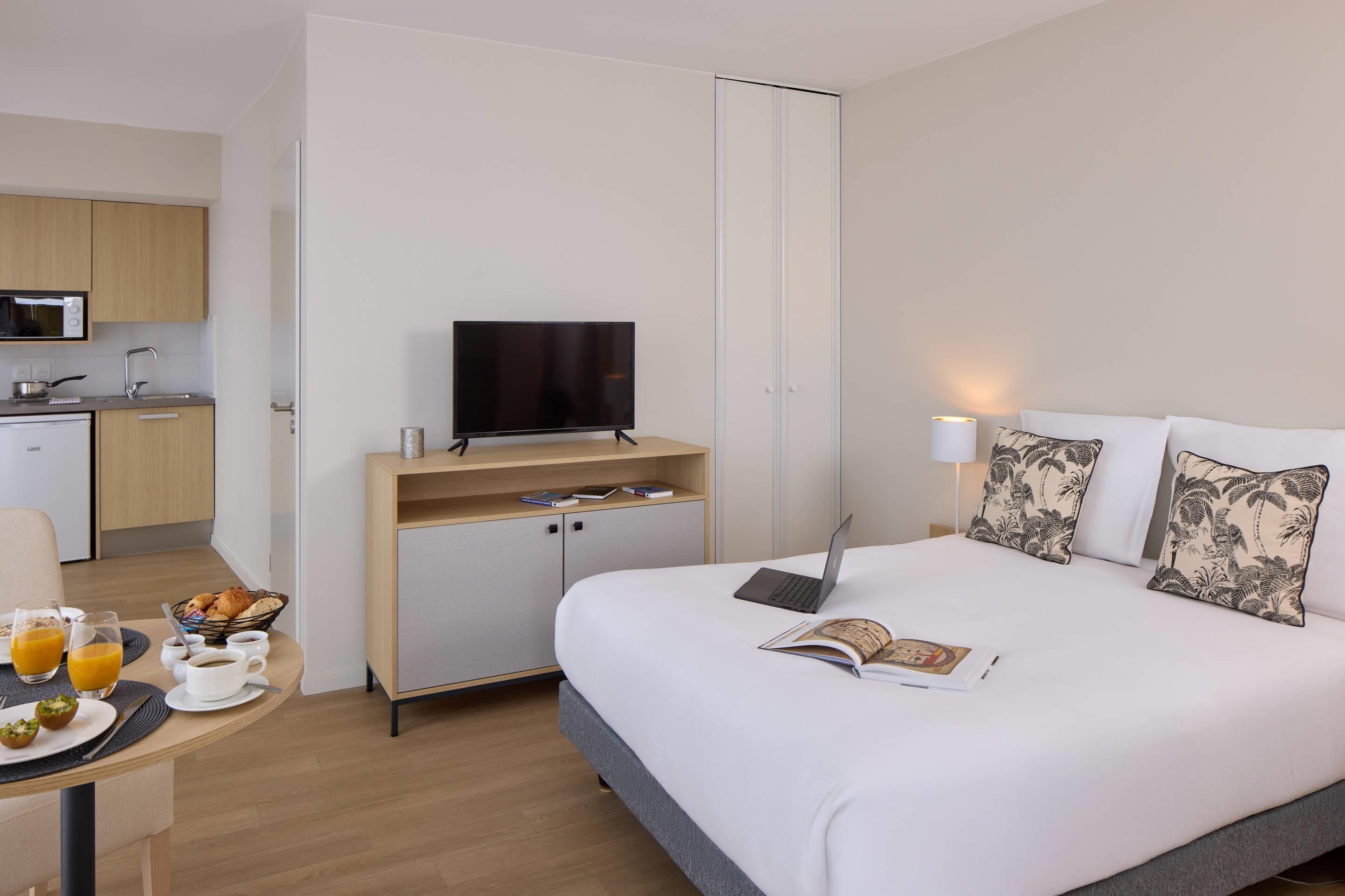 A hotel room with a bed, a desk with a laptop, a television, a dining table with breakfast, and a kitchen area.