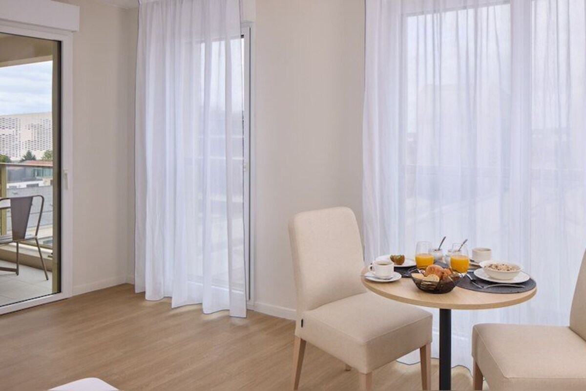Een kamer met een ronde tafel gedekt voor het ontbijt, twee stoelen, een balkon met een stoel en dunne gordijnen.