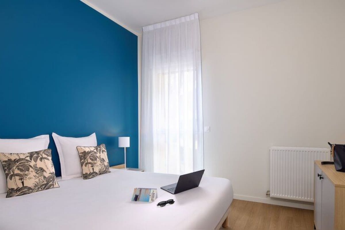 Habitación de hotel con cama, ordenador portátil, gafas de sol, bolso negro y radiador.
