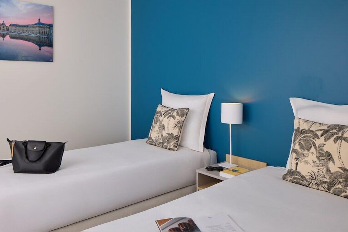 Habitación de hotel con dos camas, una mesita de noche, una lámpara, un bolso de mano y un cuadro de un edificio.