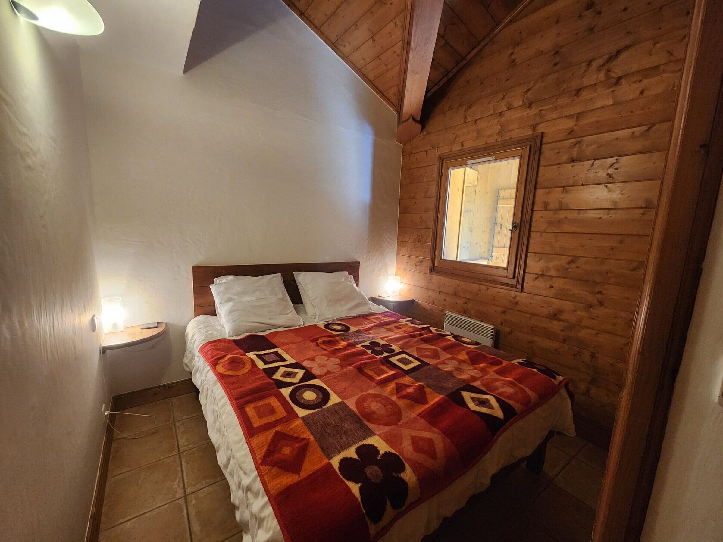 A cozy room with a wooden ceiling, a bed with a patterned blanket, a nightstand with a lamp, and a window with a view.