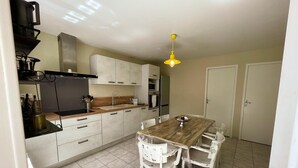 A kitchen with a wooden dining table and white chairs, a yellow hanging light fixture, and a tiled floor. - Family cottage with private terrace near Beauval (Meusnes)