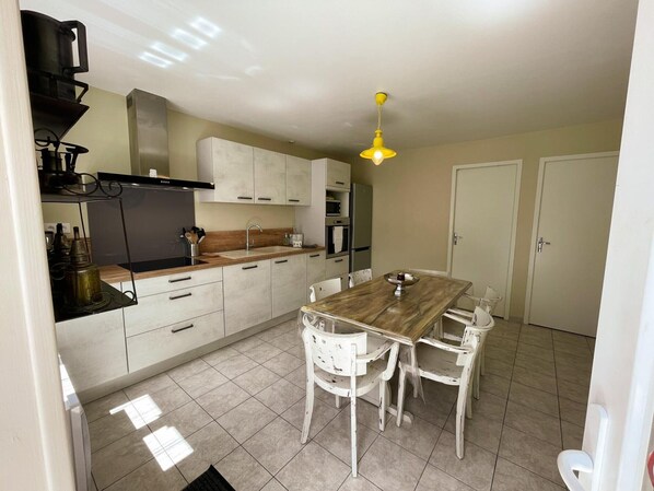 A kitchen with a wooden dining table and white chairs, a yellow hanging light fixture, and a tiled floor.