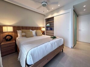 A modern bedroom with a wooden headboard, a large bed with white bedding, two bedside lamps, a nightstand, a built-in wardrobe, and a ceiling fan. - Beautiful new apartment steps from the beach (Punta Cana)