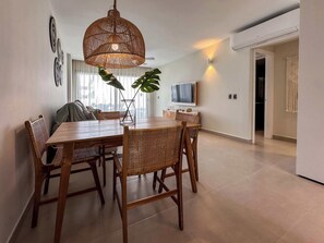 A dining area with a wooden table and chairs, a woven pendant light, and a large window with curtains. - Beautiful new apartment steps from the beach (Punta Cana)