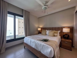 A modern bedroom with a large bed, wooden headboard, bedside lamps, and a view of a balcony.