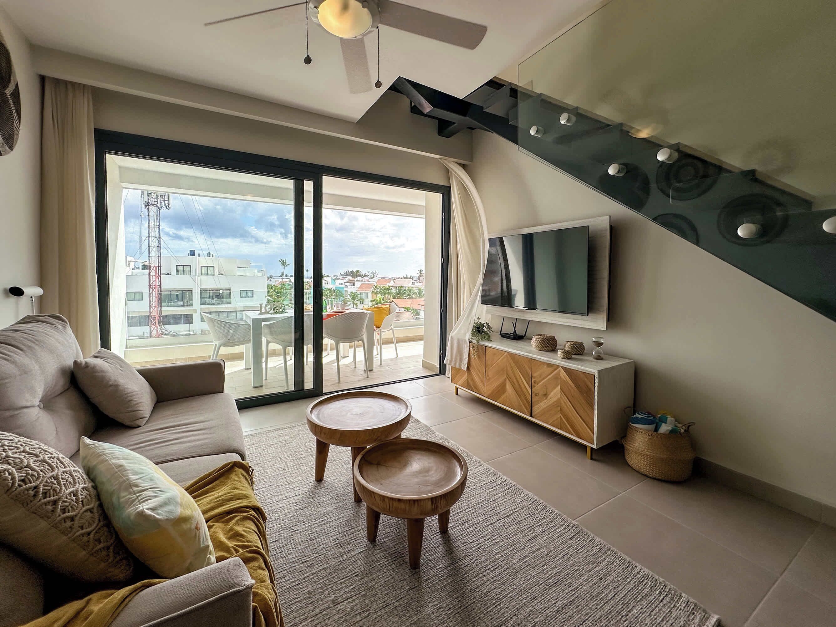 Uma sala de estar moderna com um sofá, duas mesas laterais de madeira, uma TV de tela plana e uma escada.
