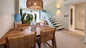 Et moderne spiseområde med et træbord, fletstole, en glas trappe og en hængende lampe.