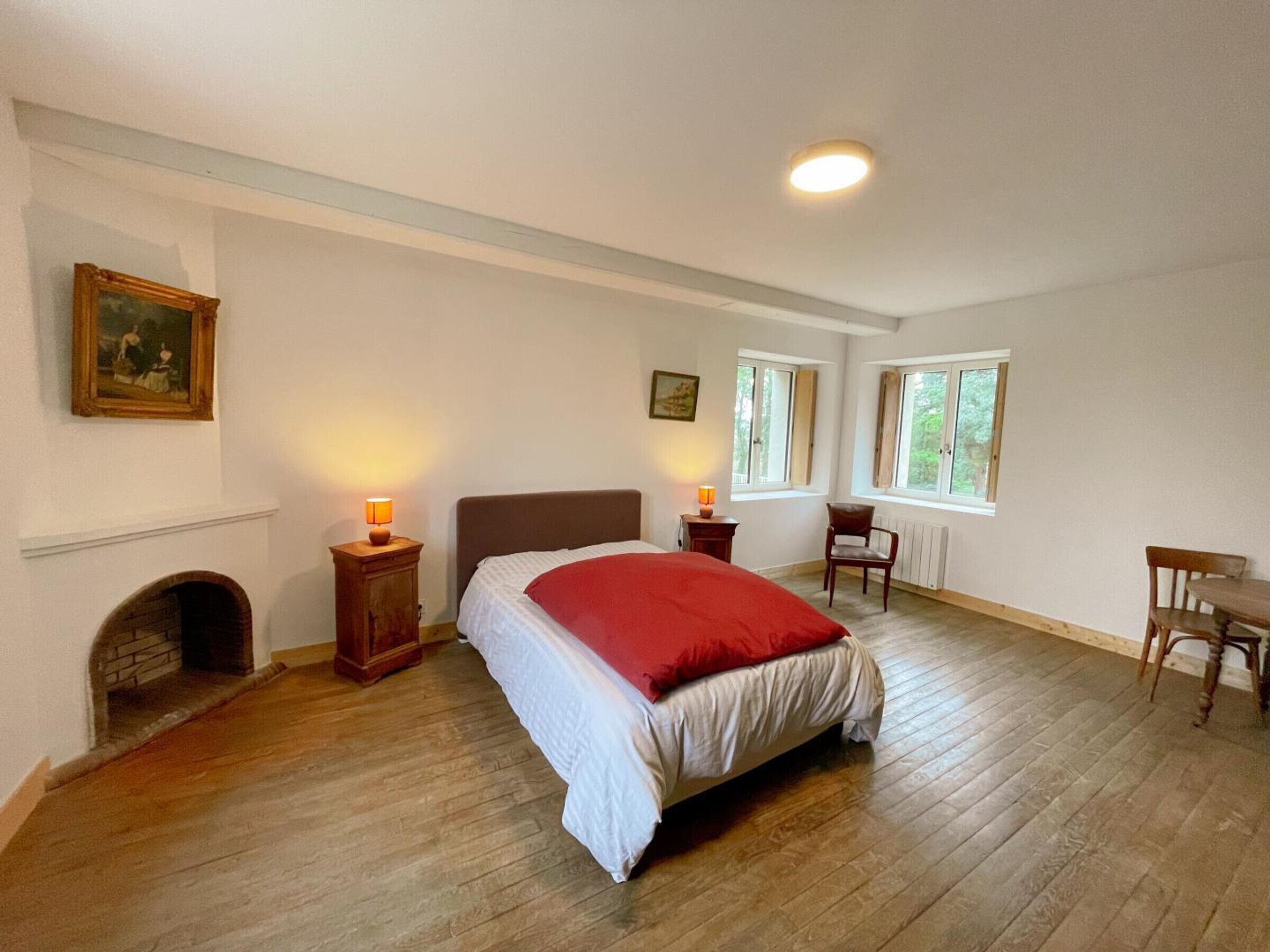 A bedroom with a bed, two bedside tables with lamps, a fireplace, and two windows.