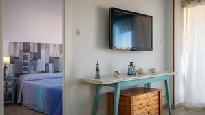 Ein Schlafzimmer mit einem Bett, einem Nachttisch mit Lampe, einem am Wand montierten Fernseher und einem Fenster mit Vorhang.