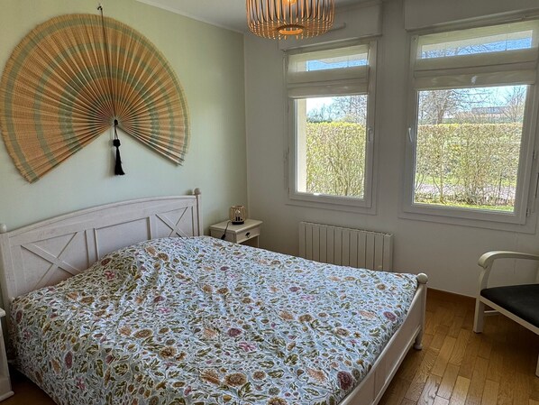 Et soveværelse med en seng, et natbord, en lampe, en stol og to vinduer.